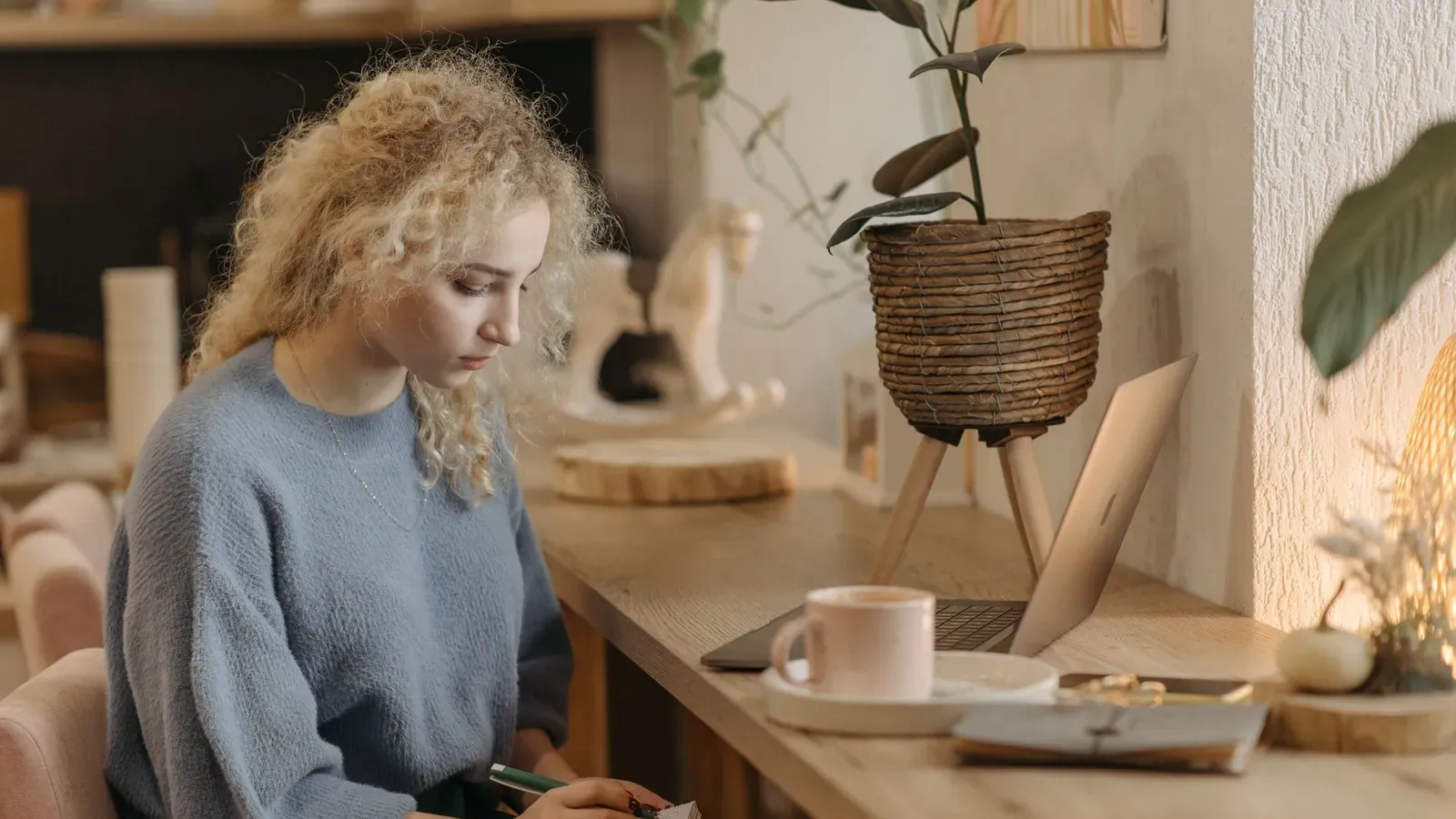 Kvinna skriver i ett block vid ett träskrivbord med bärbar dator och krukväxter