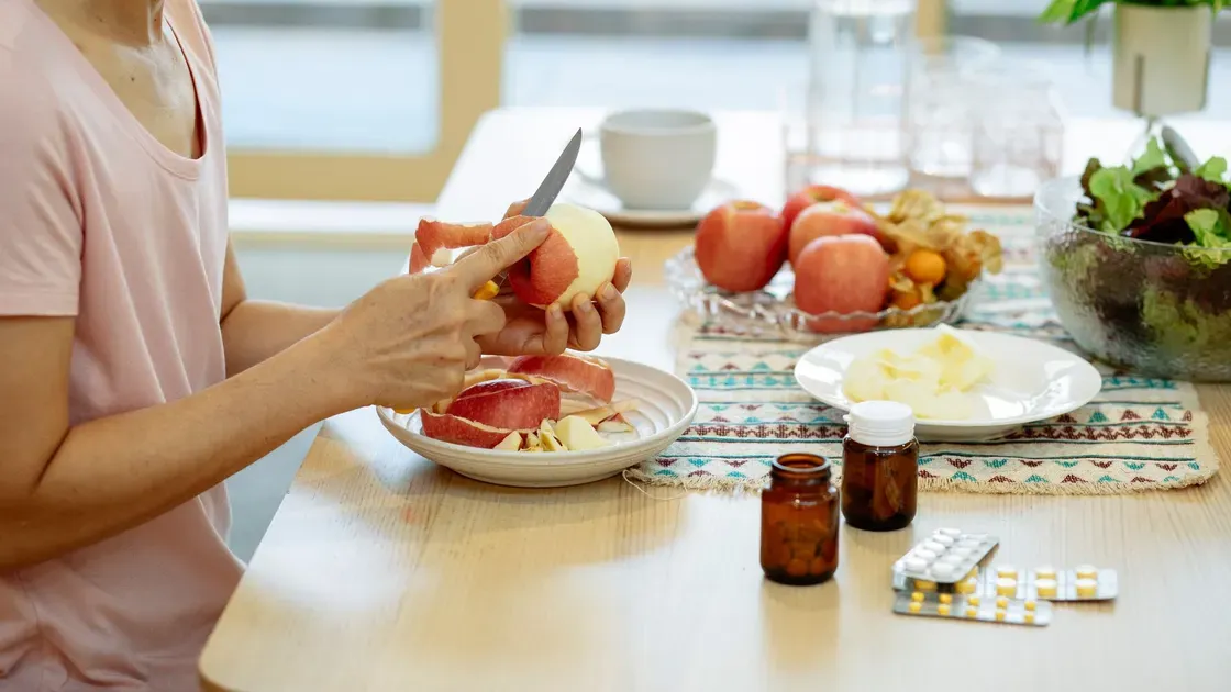 Kvinna skalar ett äpple vid ett köksbord där medicinburkar och tablettkartor står framme