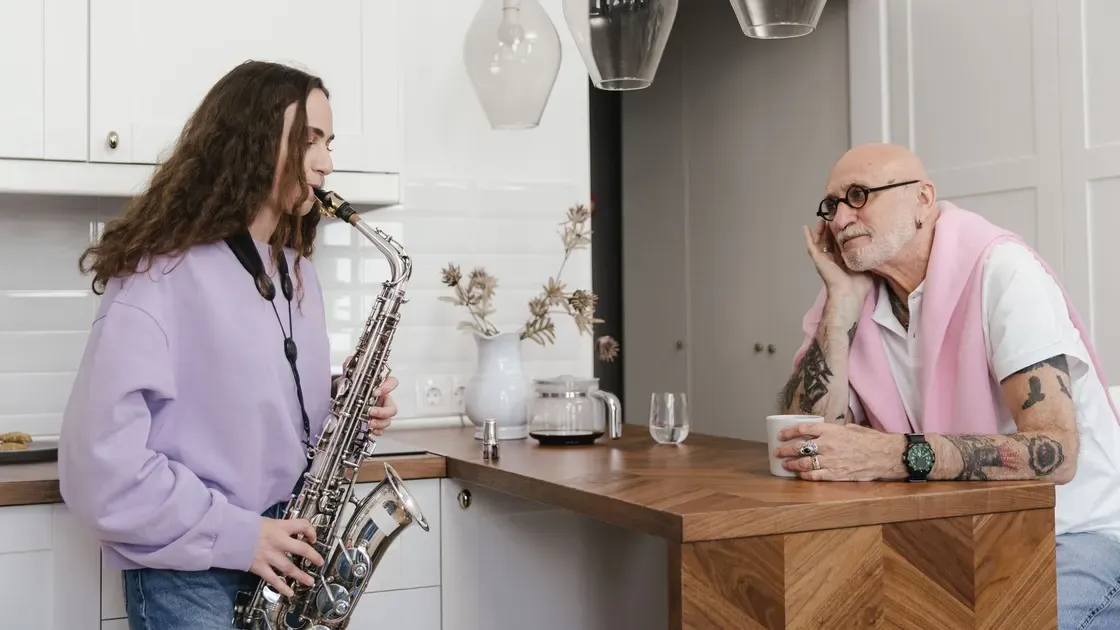 Ung person spelar saxofon i ett kök medan en äldre man lyssnar vid köksön