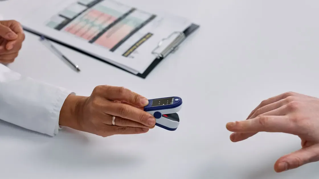 Vårdpersonal i vit rock räcker fram en blå pulsoximeter över ett skrivbord till en utsträckt hand