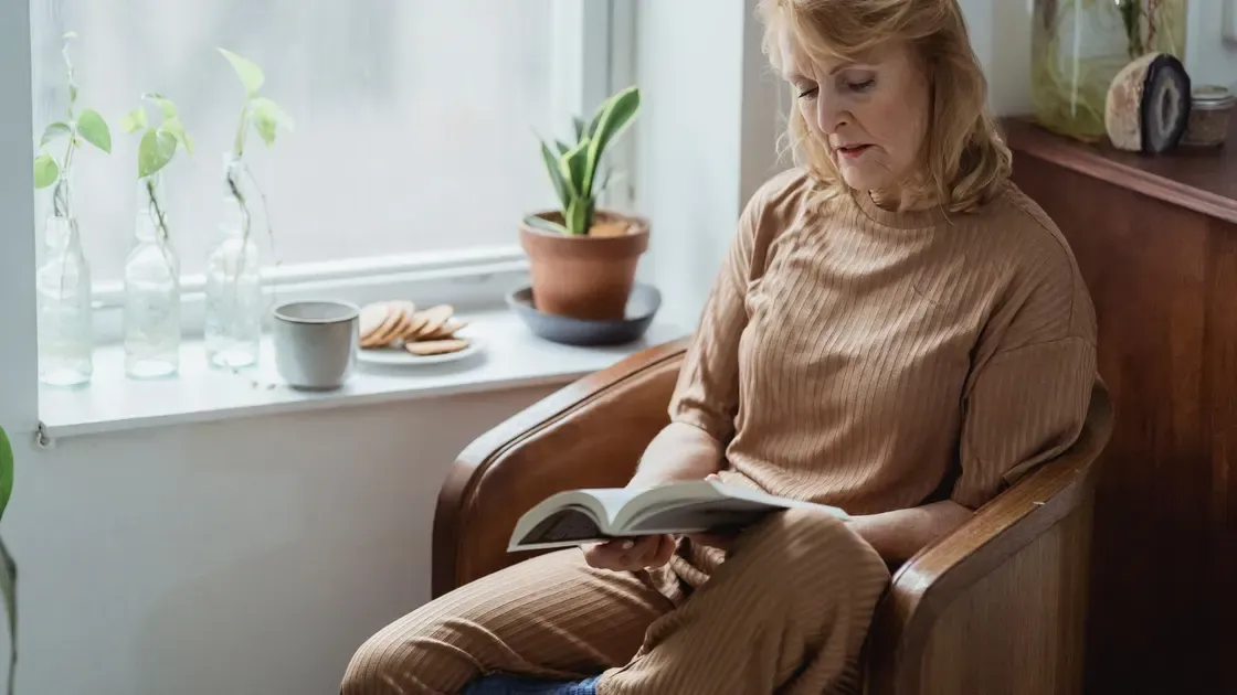 Kvinna sitter i en liten träfåtölj vid ett fönster och läser en bok
