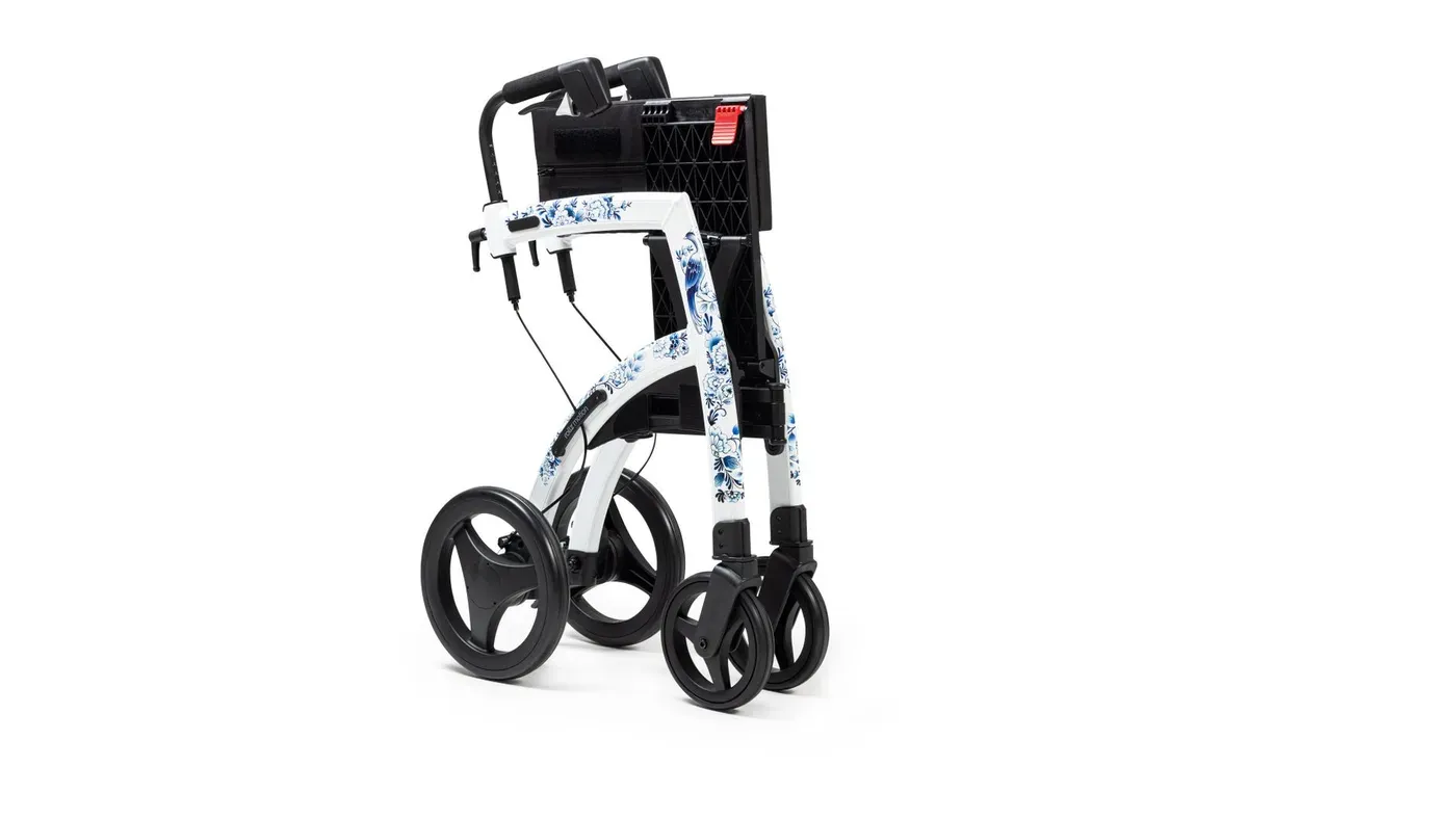 Vit rollator med blommönstrad ram, stora bakhjul och svart korg, sedd snett bakifrån