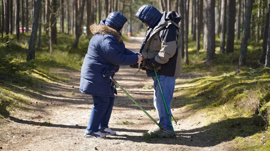 Två personer justerar handremmarna på sina gåstavar på en skogsstig