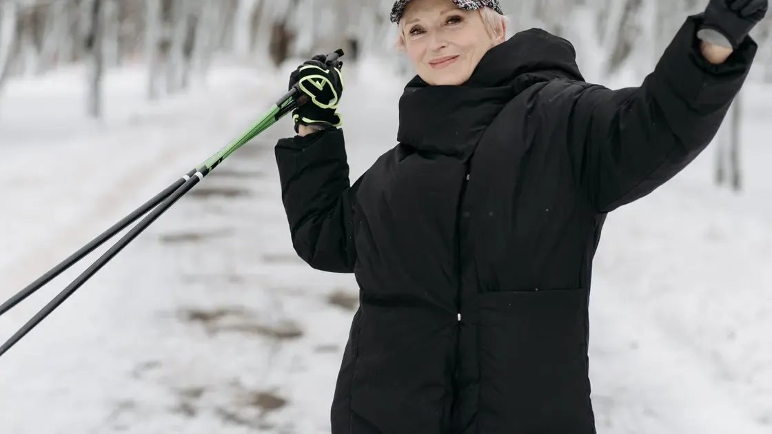 Äldre kvinna i svart vinterjacka och handskar håller ett par gåstavar på en snöig väg