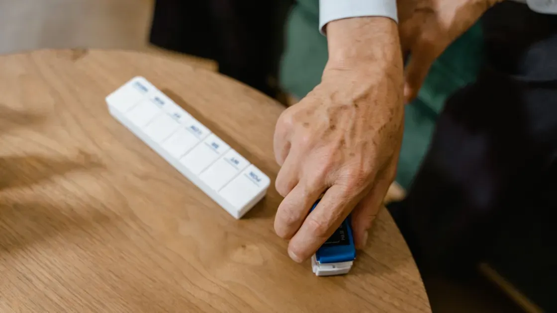 Vit veckodosett med sju fack märkta med veckodagar på ett runt träbord bredvid en hand