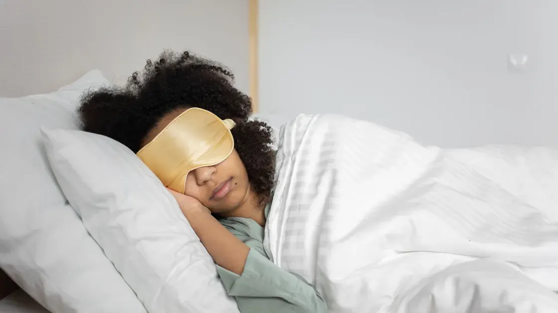 Kvinna sover på sidan med en gul sovmask i satin och kinden mot kudden