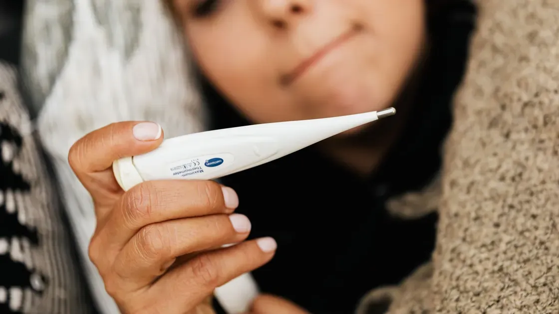Närbild på en vit digital sticktermometer som hålls i handen av en person som vilar under en filt