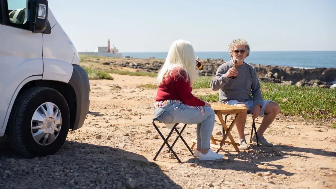 Par som sitter på hopfällbara pallar vid ett litet bord på grus intill en husbil