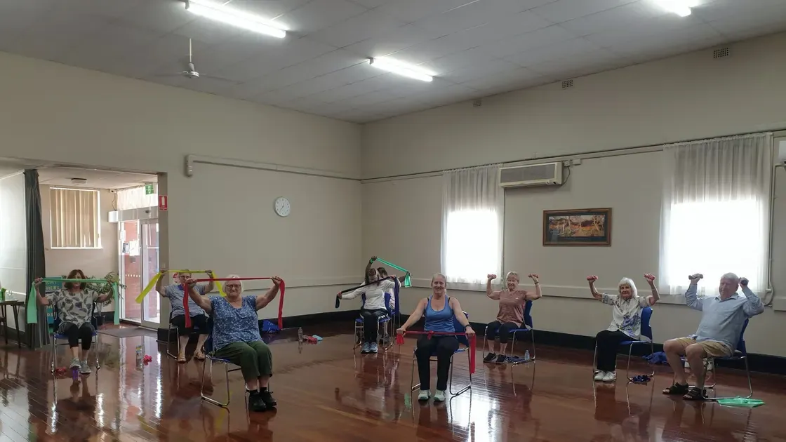 Träningsgrupp för äldre sitter på stolar i en gymnastiksal och tränar med gummiband
