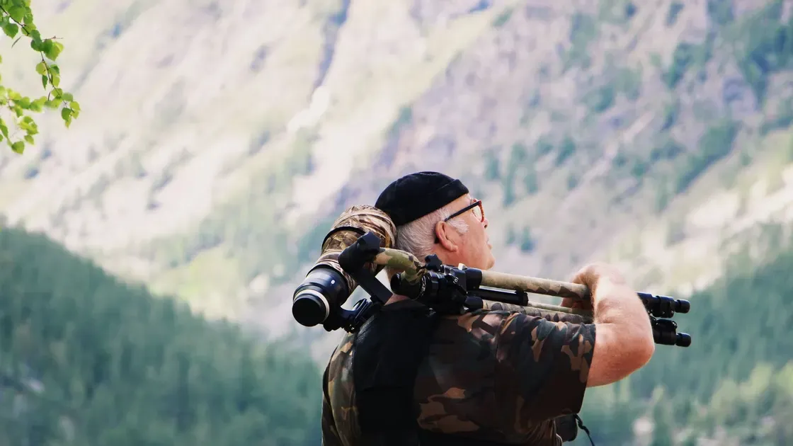 Äldre man som bär ett stativ med optik över axeln i bergen