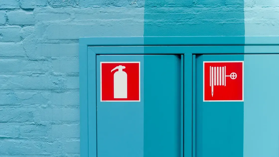Två röda brandskyltar på ett turkost skåp, den ena med en brandsläckare och den andra med en brandslang