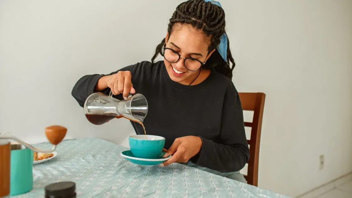 Kvinna med glasögon häller kaffe ur en liten glaskanna i en turkos kopp vid frukostbordet