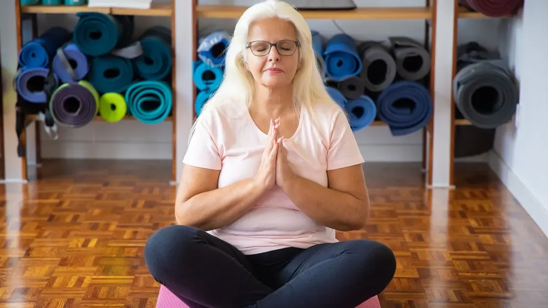 Kvinna med glasögon sitter med korsade ben på en rosa yogamatta framför en hylla med ihoprullade träningsmattor