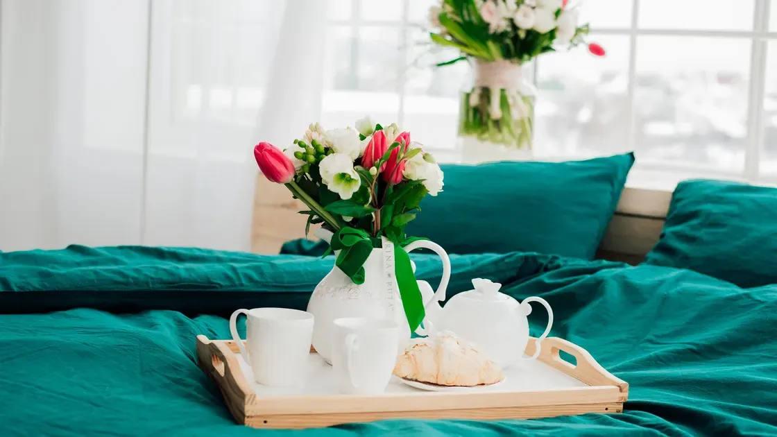 Sängbricka i trä med kaffekoppar, kanna och blommor på en bäddad säng