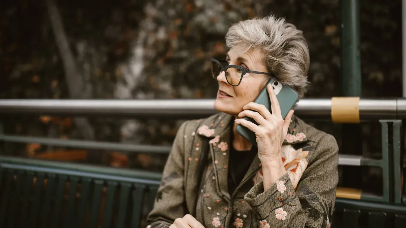 Äldre kvinna med glasögon som talar i mobiltelefon på en bänk utomhus