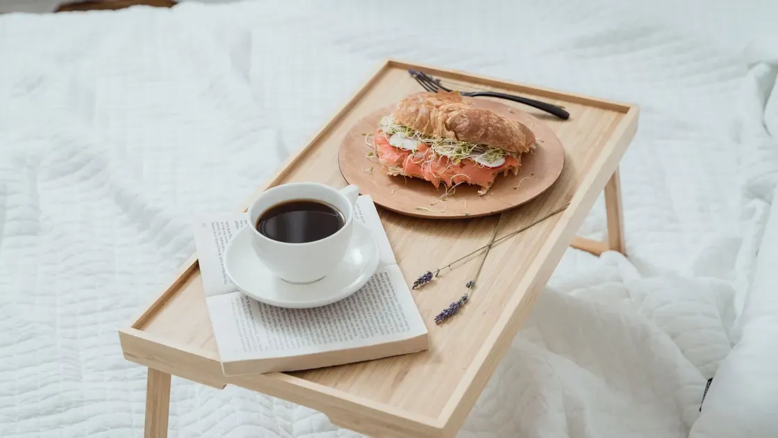 Sängbricka i trä med kaffekopp, uppslagen bok och en smörgås på en vit säng