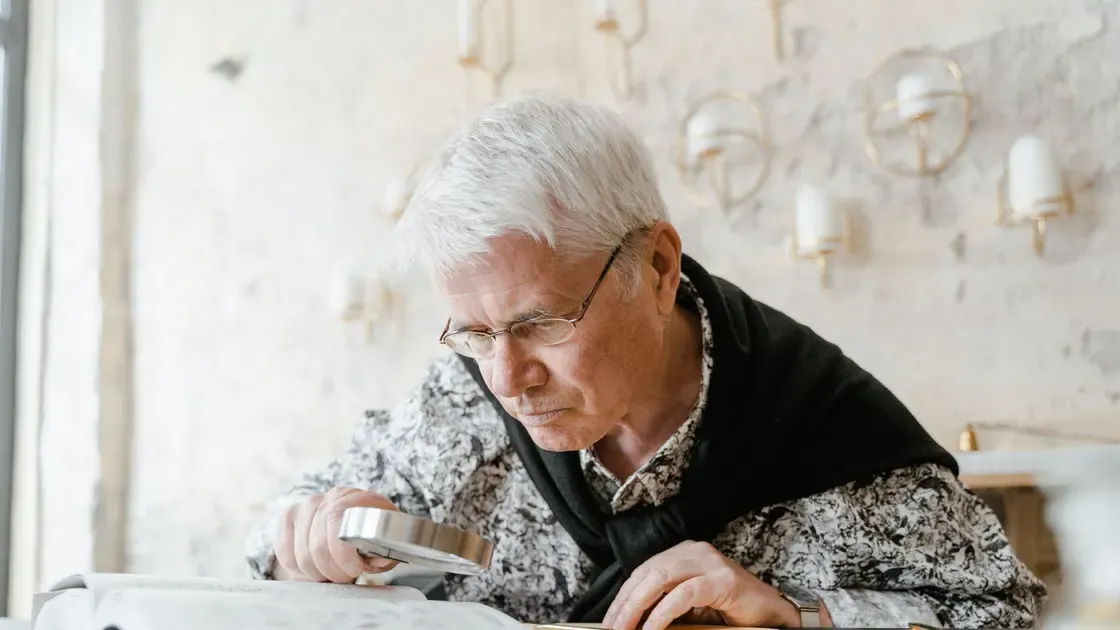 Äldre man med läsglasögon lutar sig över ett papper och håller ett förstoringsglas nära texten