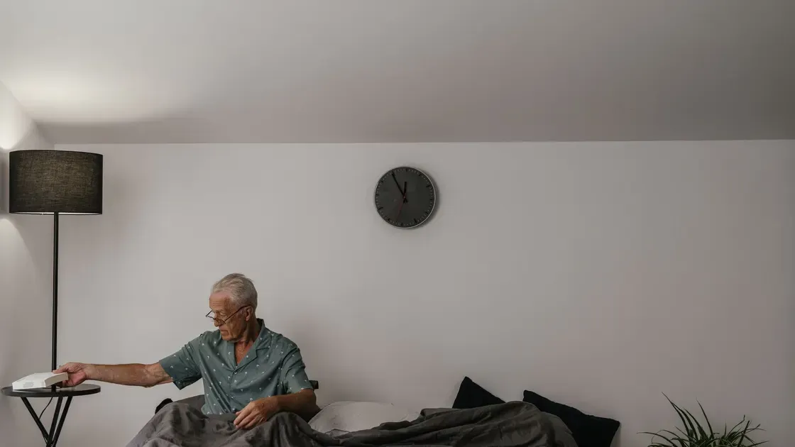 Man i pyjamas sitter på sängkanten och sträcker sig efter en vit väckarklocka på bordet vid golvlampan
