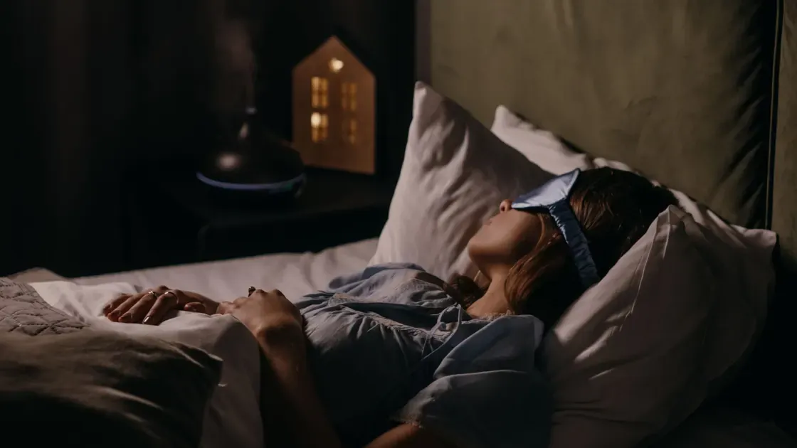 Kvinna sover på rygg med ljusblå sovmask i ett mörkt sovrum med tänd nattlampa