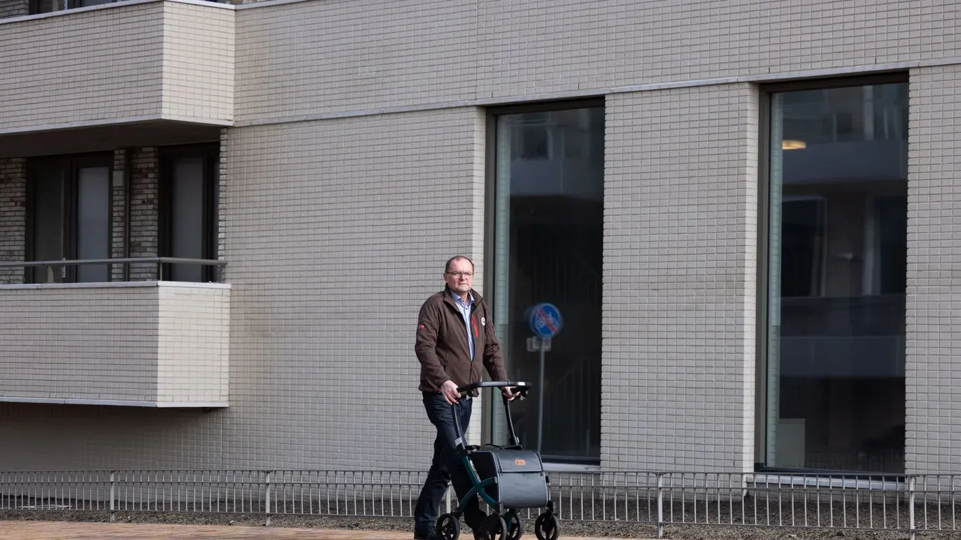Man i brun jacka går med en rollator med väska längs en husfasad
