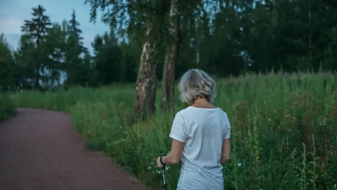 Äldre kvinna sedd bakifrån går med gåstav på en grusstig kantad av högt gräs
